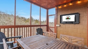 Eine Holzterrasse mit Tisch, Stühlen, einem an der Wand befestigten Fernseher und Blick ins Freie.