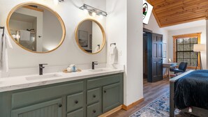 Ein Badezimmer mit zwei Waschbecken, einem Spiegel und einem Schminktisch mit Schubladen. Im angrenzenden Zimmer steht ein Bett mit dunkler Bettdecke.