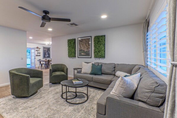 A living room with a grey sofa, two armchairs, a coffee table, and a ceiling fan. There are framed pictures on the wall and a window with blinds.