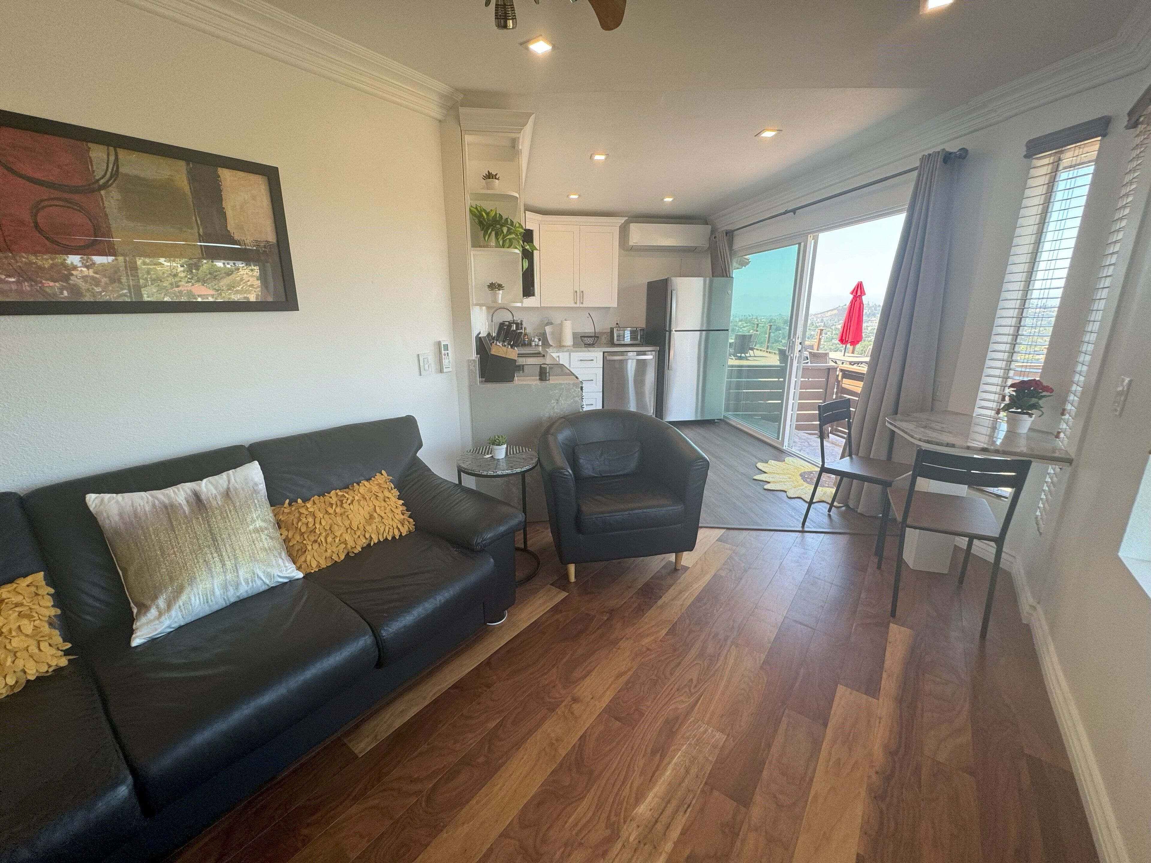 Un salon moderne avec un canapé en cuir noir, un petit fauteuil noir et un coin repas comprenant une table et des chaises. On aperçoit, au fond, une cuisine avec des meubles blancs et un réfrigérateur.