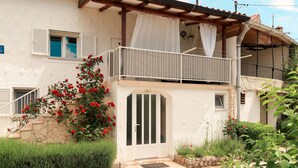 Une maison à deux étages avec un balcon, un escalier en pierre et un jardin agrémenté de fleurs rouges.