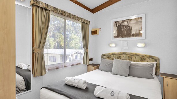 A hotel room with a large bed, a headboard with a patterned design, two bedside tables, a window with curtains, and a framed picture on the wall.