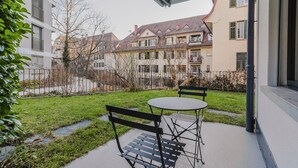 En udendørs terrasse med et bord og to bænke, omgivet af en velplejet græsplæne og et hegn.