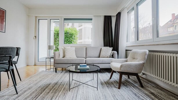 En moderne stue med en grå sofa, et rundt sofabord og en hvid lænestol. Der er et stort vindue med gardiner, en radiator og udsigt til bygninger.