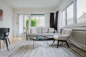 A modern living room with a grey sofa, a round coffee table, and a white armchair. There is a large window with curtains, a radiator, and a view of buildings outside. - Blueground | Unterstrass, balcony & washer (Zurich)