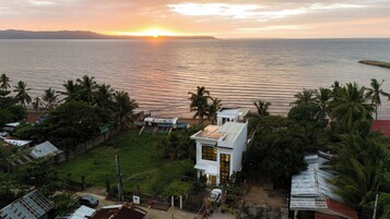 Vista costera con un atardecer sobre el mar, un edificio blanco con puerta amarilla y palmeras.