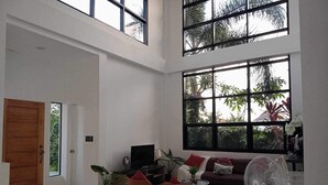 Een ruime woonkamer met een hoge plafond, grote ramen, een bank, een televisie en een ventilator.