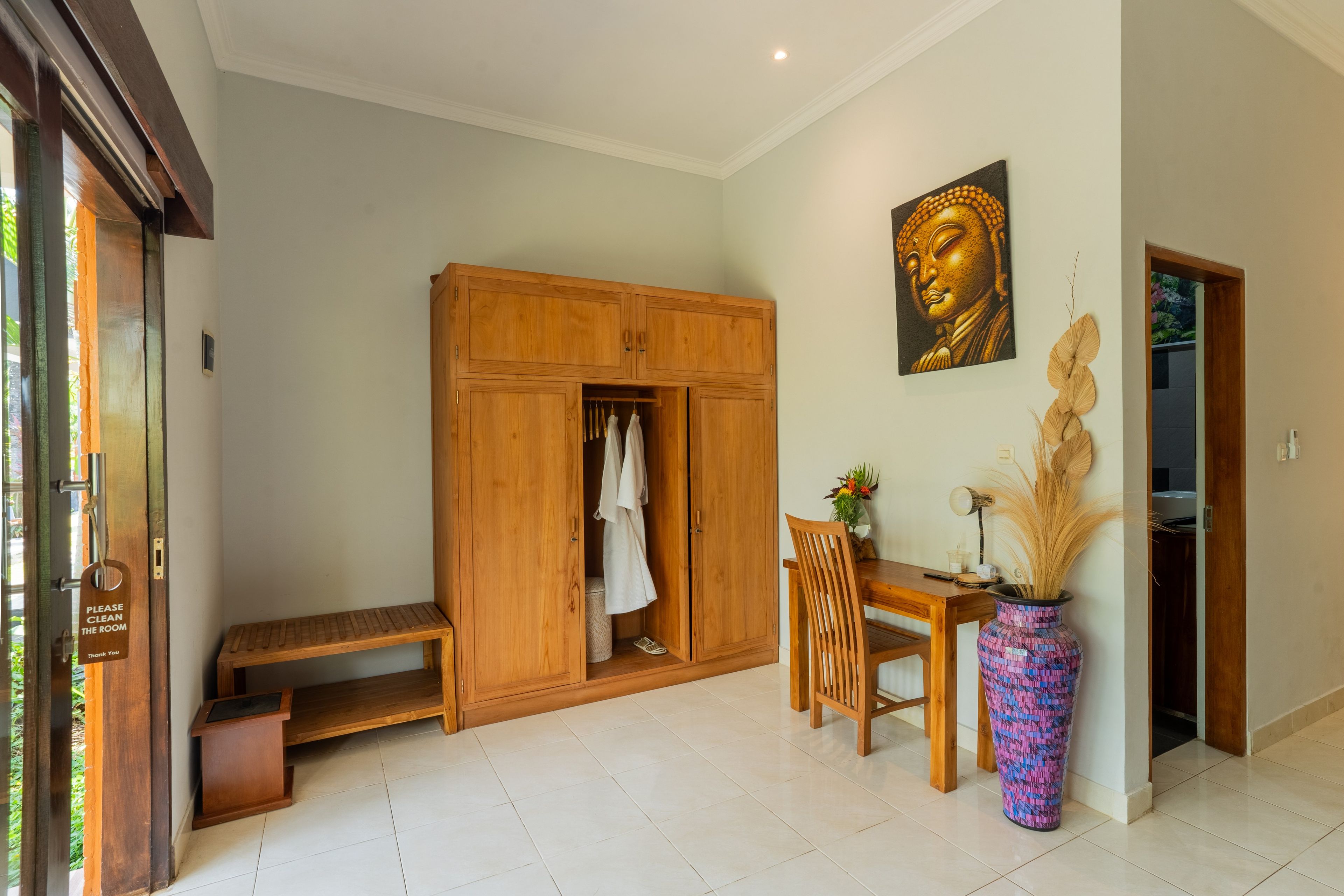 Um quarto com guarda-roupa de madeira, um banco, uma mesa de jantar com cadeiras, um vaso com plantas secas e uma estátua de Buda na parede.