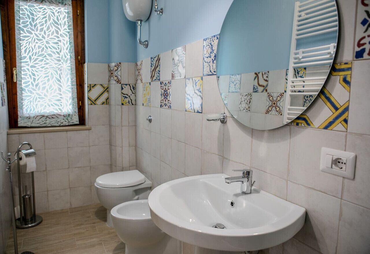 A bathroom with a white sink, two toilets, patterned tiles, a mirror, and a window with a decorative screen.