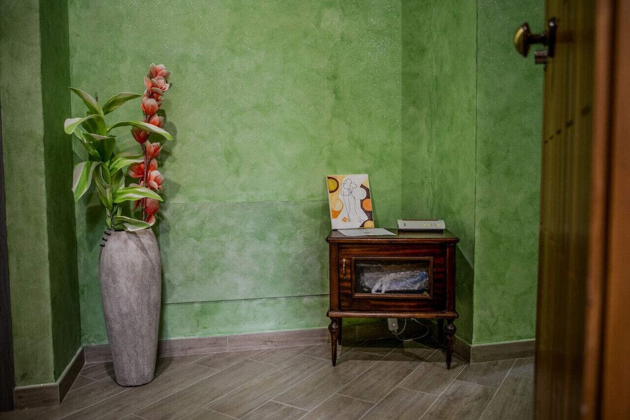 Et hjørne med en grøn væg med struktur, en høj vase med røde blomster og et lille træsidebord med et indrammet billede og en bog.