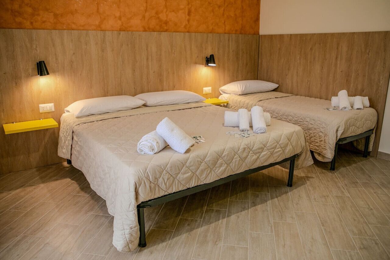 A hotel room with two beds, each with a beige quilted bedspread and white pillows. There are two bedside tables with lamps, and towels neatly folded on the beds.
