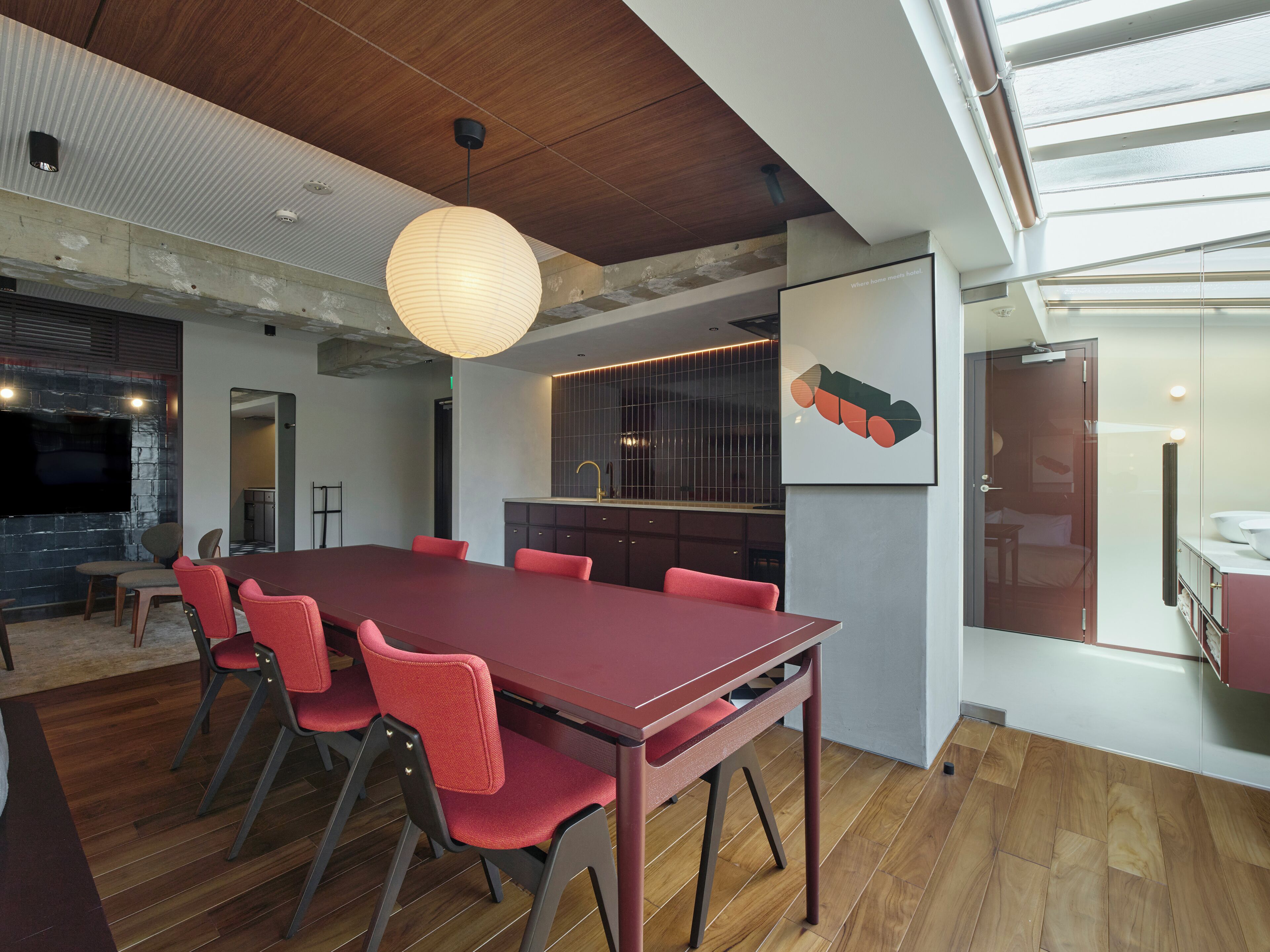 Habitación de hotel moderna con una gran mesa de comedor de madera, sillas rojas, una zona de cocina y un amplio ventanal que proporciona luz natural.
