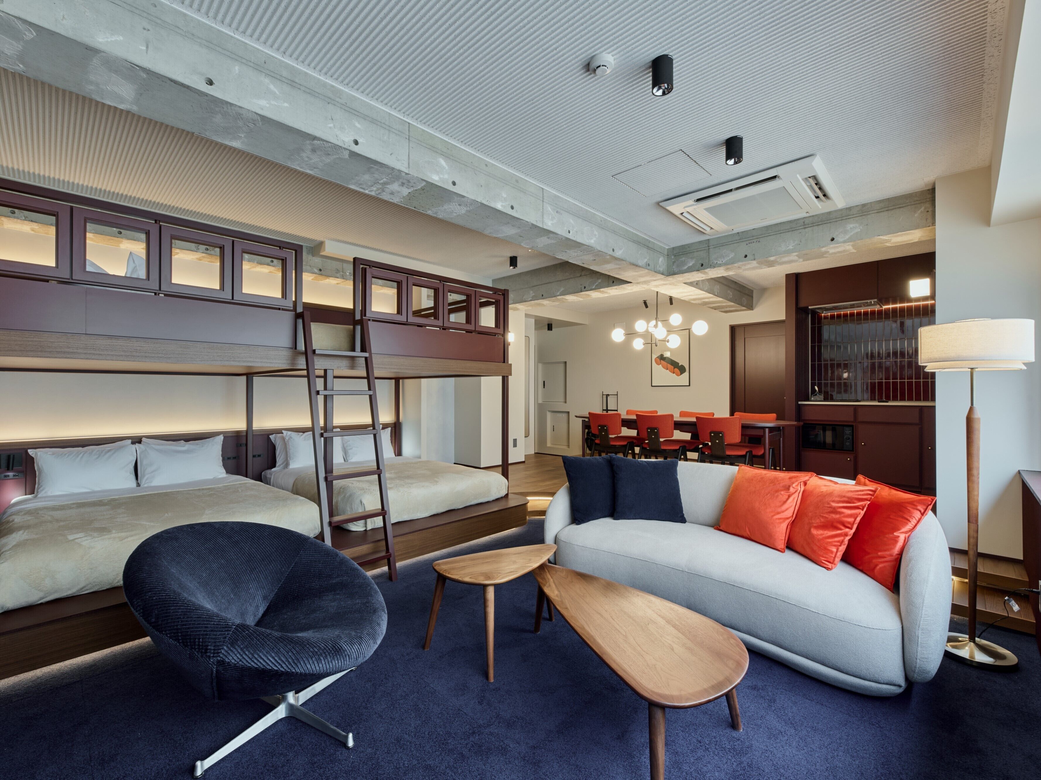 A modern hotel room with a bunk bed, a sofa, a chair, a small table, and a dining area with chairs and a chandelier.