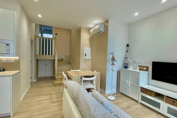 Una sala de estar moderna con un sofá, una mesa de comedor y un televisor. Una escalera conduce a un nivel superior.