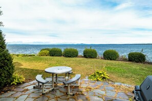 Un patio con tavolo e sedie affacciato su un lago, circondato da cespugli curati e un sentiero in pietra.