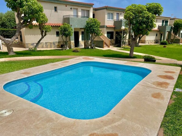 Piscina retangular com borda azulejada, rodeada por um deck de concreto. Árvores e um edifício com telhado em azulejos aparecem ao fundo.