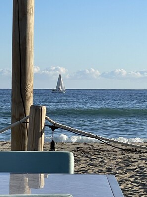 Une scène de plage avec un voilier à l’horizon, des poteaux en bois et une chaise à laquelle est attachée une corde.