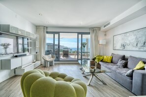 Un salon moderne doté d’une grande porte vitrée donnant sur un balcon, d’un canapé gris, d’un fauteuil vert et d’un téléviseur à écran plat.