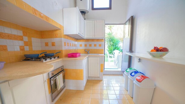 A compact kitchen with a gas stove, oven, and a sink. There are white cabinets and a tiled backsplash. A window provides natural light, and a bowl of fruit is on a shelf. - Marisole House With Sea View, Santa Cesarea Terme, Italy (Santa Cesarea Terme)