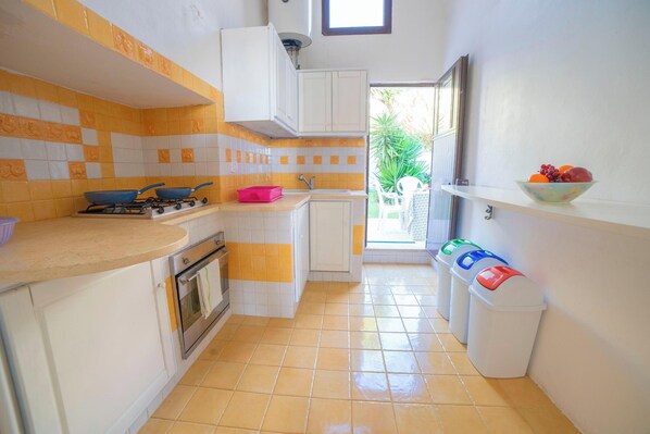A compact kitchen with a gas stove, oven, and a sink. There are white cabinets and a tiled backsplash. A window provides natural light, and a bowl of fruit is on a shelf. - Marisole House With Sea View, Santa Cesarea Terme, Italy (Santa Cesarea Terme)