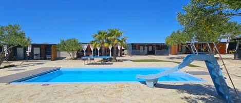 A pool area with a clear blue sky, stone flooring, and modern buildings in the background.
