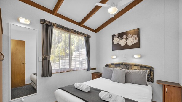 A bedroom with a large bed, a wooden headboard, a window with curtains, a painting on the wall, and a bedside table. - 2 Bedroom Family Villa (Metung)