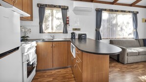 A compact kitchen with wooden cabinets, a built-in stove, and a sink. There is a small island with a toaster and a kettle. The room has a wooden ceiling and a sofa. - 2 Bedroom Family Villa (Metung)