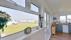 A modern kitchen with large windows, a clock, and a view of a white car and building outside. - D46 Wrangarrie, Riviere Towans (Hayle)