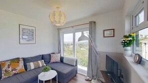 A modern living room with a grey sofa, a small white table, a TV, and a large window with a view of greenery. - D46 Wrangarrie, Riviere Towans (Hayle)