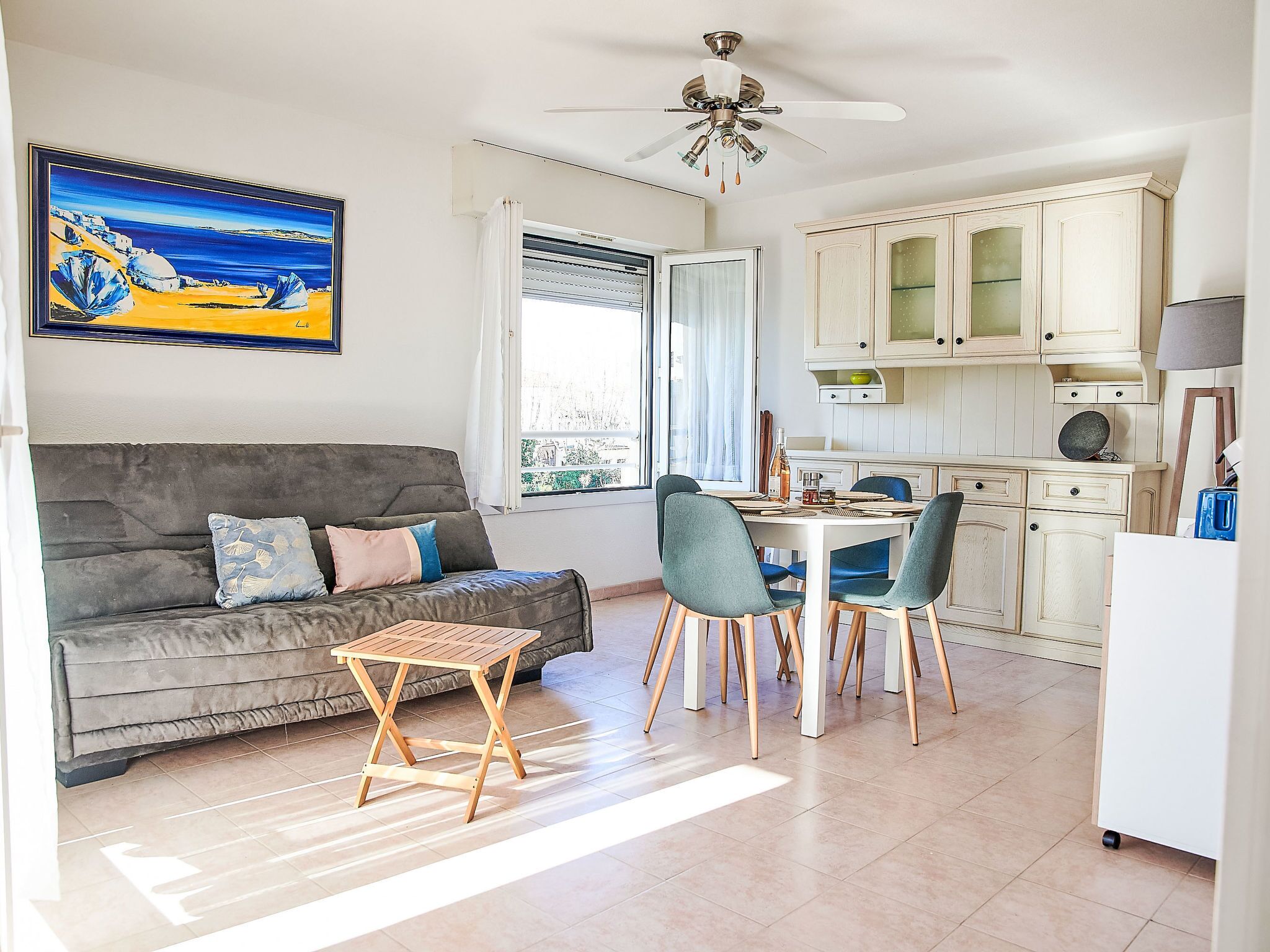 Un salon moderne avec un canapé gris, une petite table en bois et des chaises turquoise. On y trouve également un coin repas avec une table et des chaises blanches, ainsi qu'une cuisine avec des meubles blancs et un ventilateur de plafond.