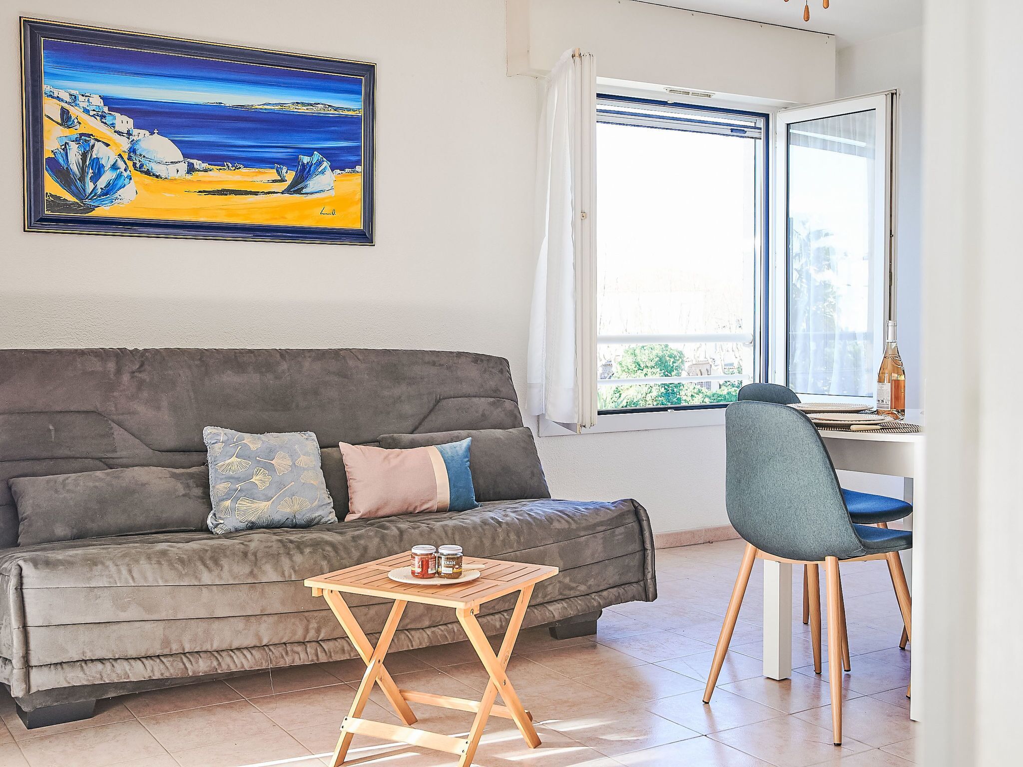 Un salon moderne avec un canapé gris, une petite table en bois et une chaise bleue. Un tableau représentant un paysage côtier est accroché au mur.