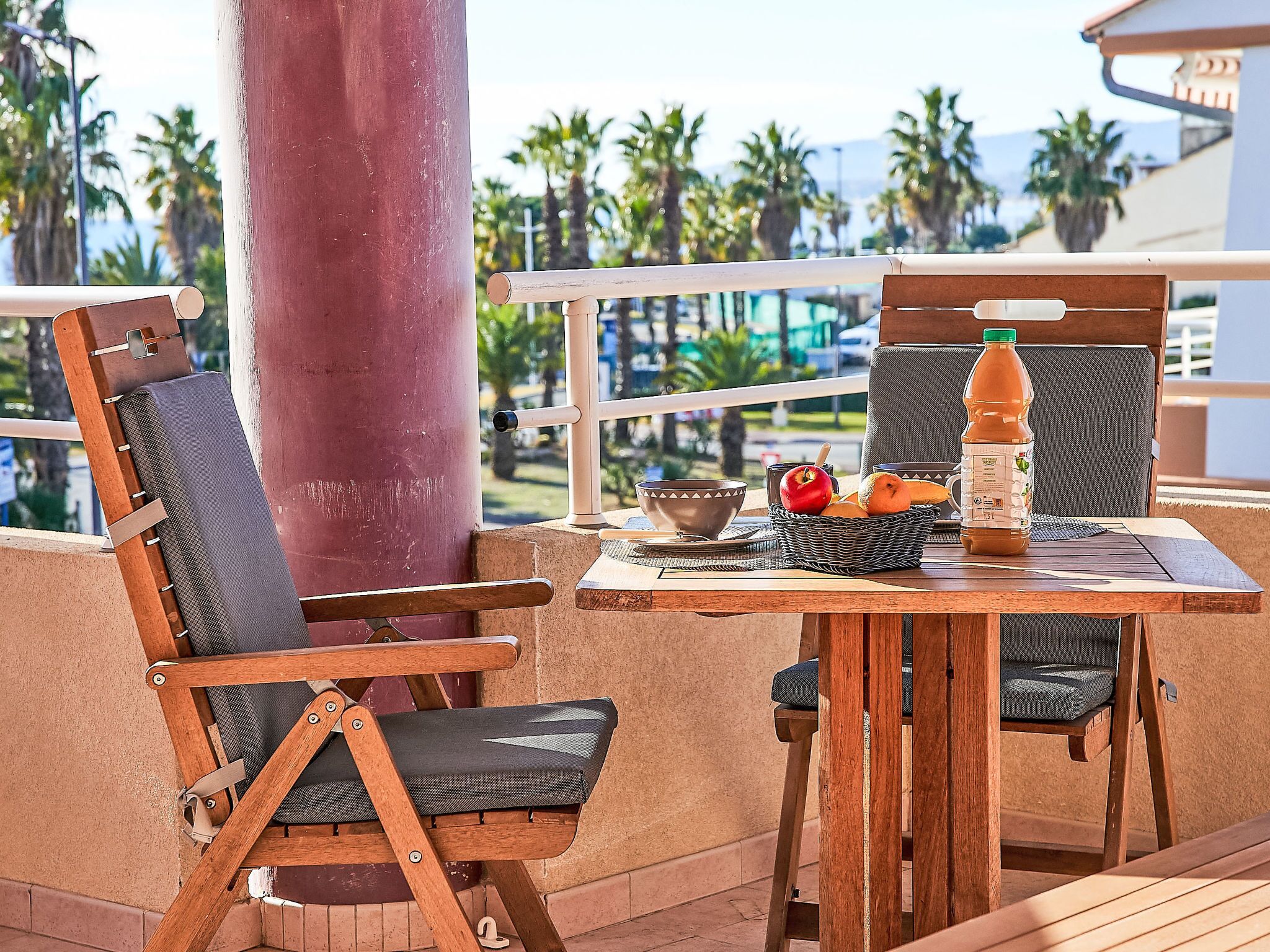 Uma mesa de madeira com uma cesta de vime contendo frutas e uma garrafa, duas cadeiras de madeira com almofadas, e uma vista de palmeiras e edifícios ao fundo.