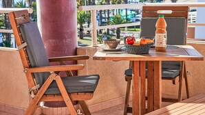 A wooden table with a wicker basket containing fruits and a bottle, two wooden chairs with cushions, and a view of palm trees and buildings in the background. - Les Rives Latines by Interhome (Fréjus)