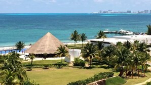 A resort with a thatched-roof structure, palm trees, and a view of the ocean and city skyline. - Beachfront 3BR - Ocean View - 70m Pool - Cancun (Cancún)