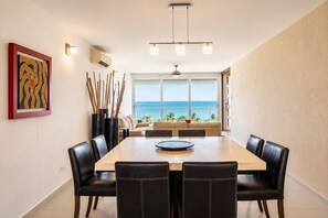 Uma área de jantar com uma mesa grande, cadeiras, um ventilador de teto e vista para o oceano.