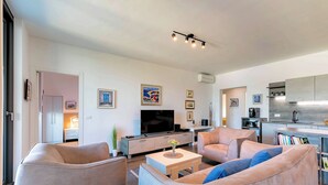 A modern living room with a sofa set, a coffee table, and a flat-screen TV. There is a kitchen area with a sink and cabinets.