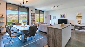 A modern living room with a dining area, large windows with a mountain view, a flat-screen TV, and a wooden coffee table.