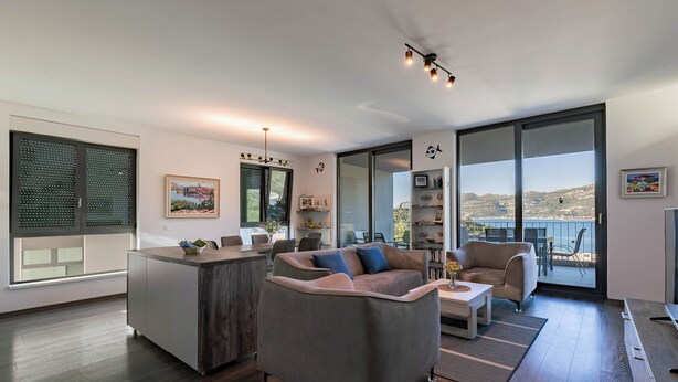A modern living room with a grey sofa, a coffee table, and a dining area. There is a large window with a view of the outdoors.