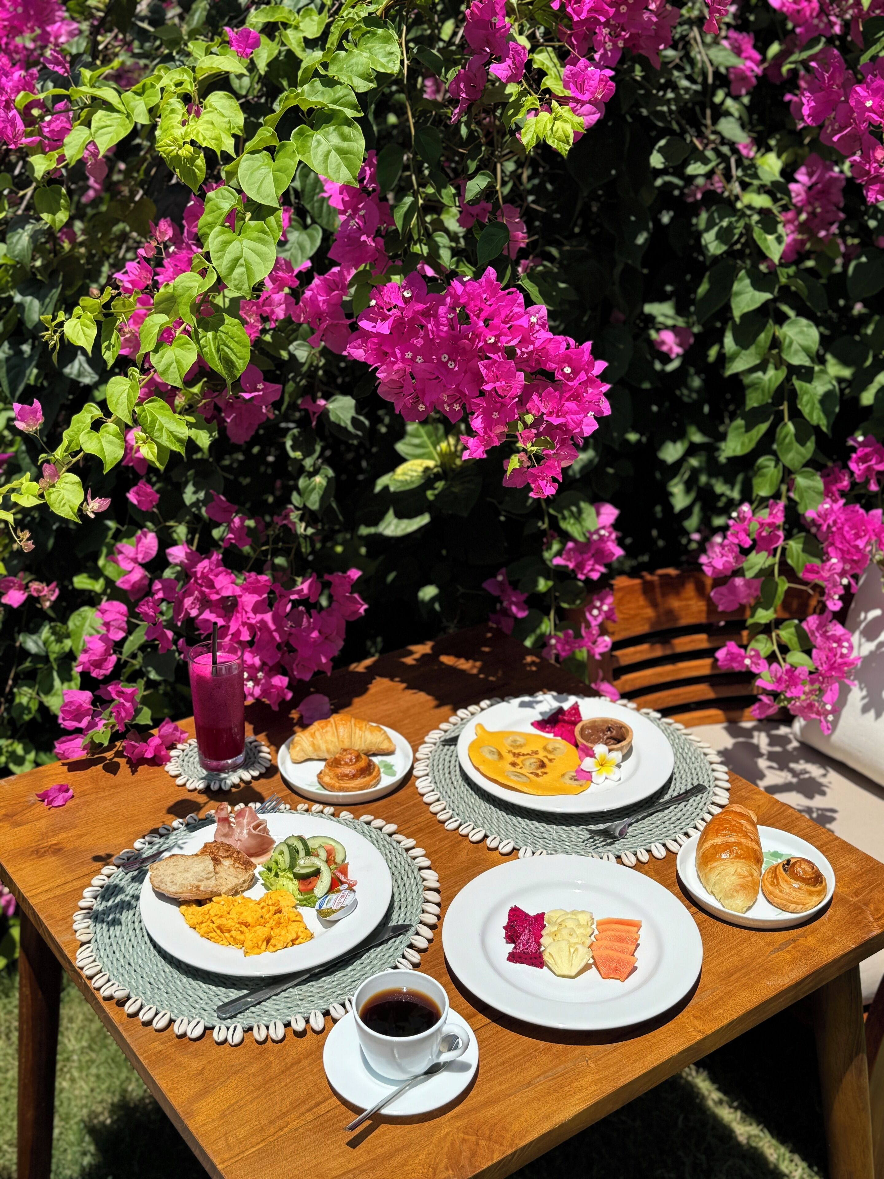 Un tavolo di legno con prodotti per la colazione, tra cui croissant, uova e una bevanda, all'aperto con vivaci fiori rosa sullo sfondo.