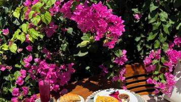 Un tavolo di legno con prodotti per la colazione, tra cui croissant, uova e una bevanda, all'aperto con vivaci fiori rosa sullo sfondo.