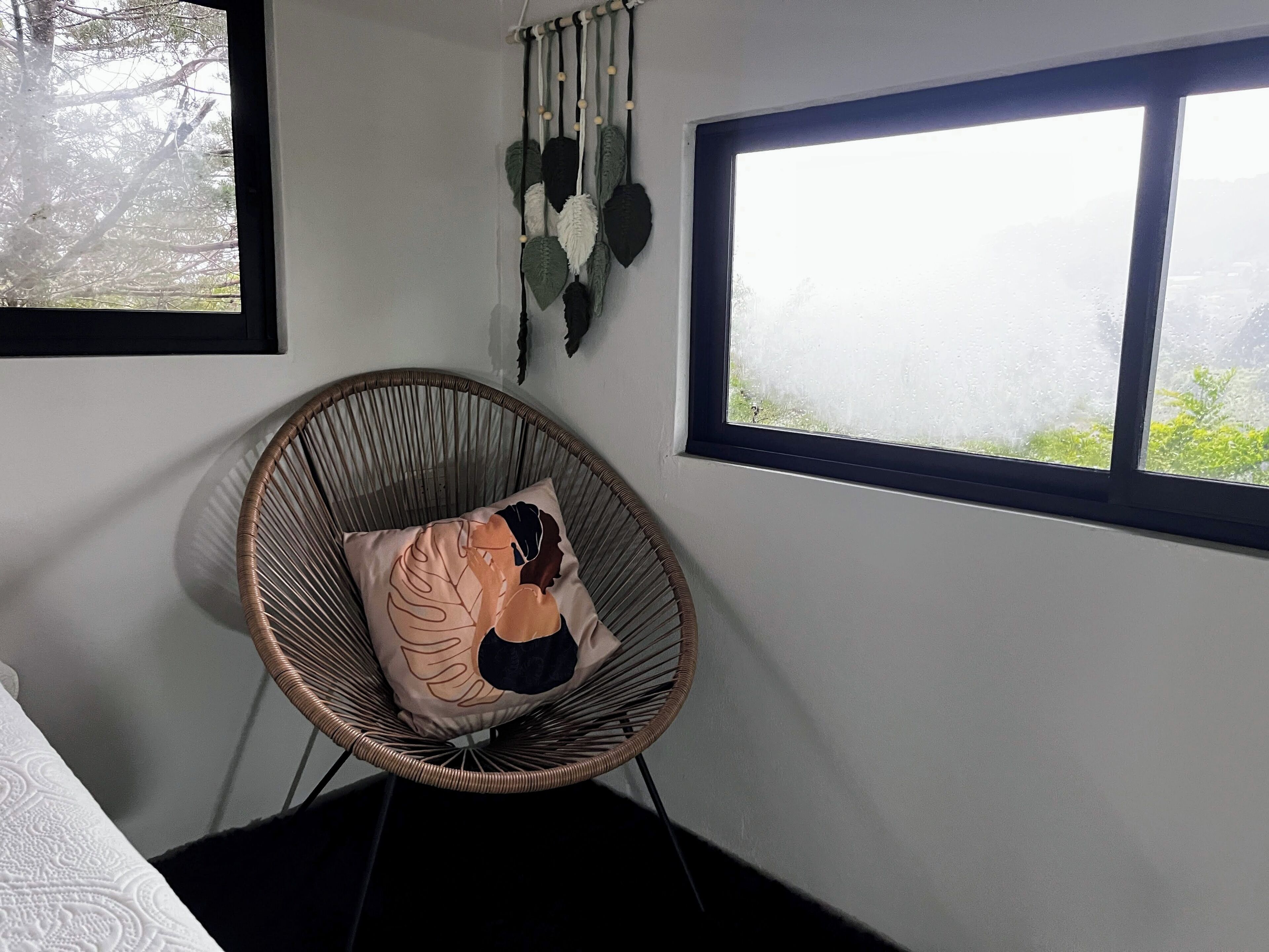A room with a wicker chair, a window with a view, and a wall-mounted macrame decoration.