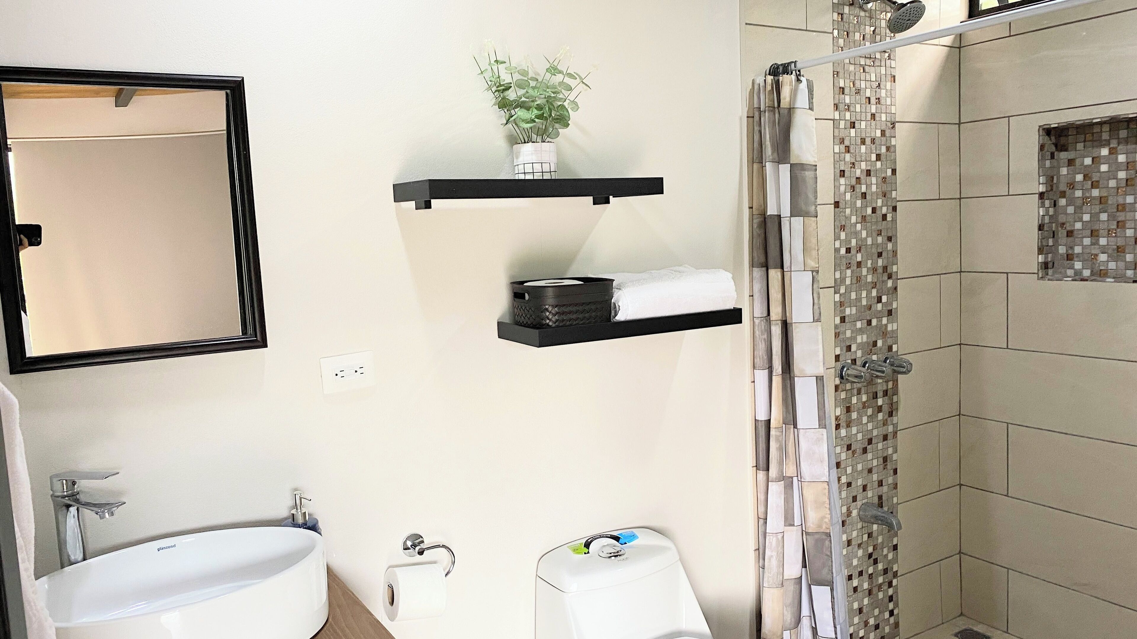 Une salle de bain moderne avec un grand miroir, un lavabo, des toilettes, une douche avec un mur recouvert de mosaïques, et deux étagères murales où sont posés une plante et une serviette.