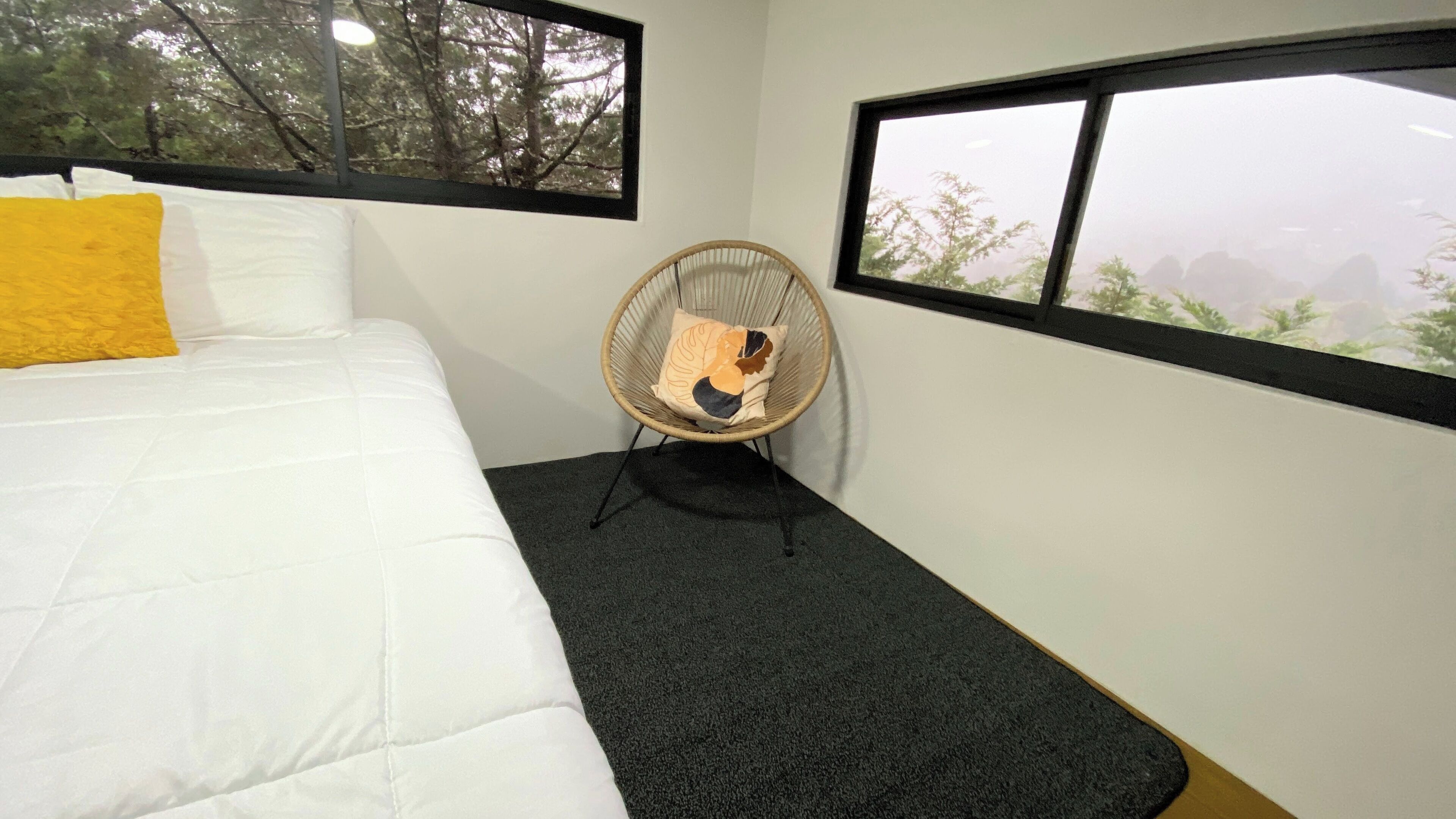 A bed with a white comforter and a yellow pillow, a wicker chair with a cushion, and a view of trees through a window.
