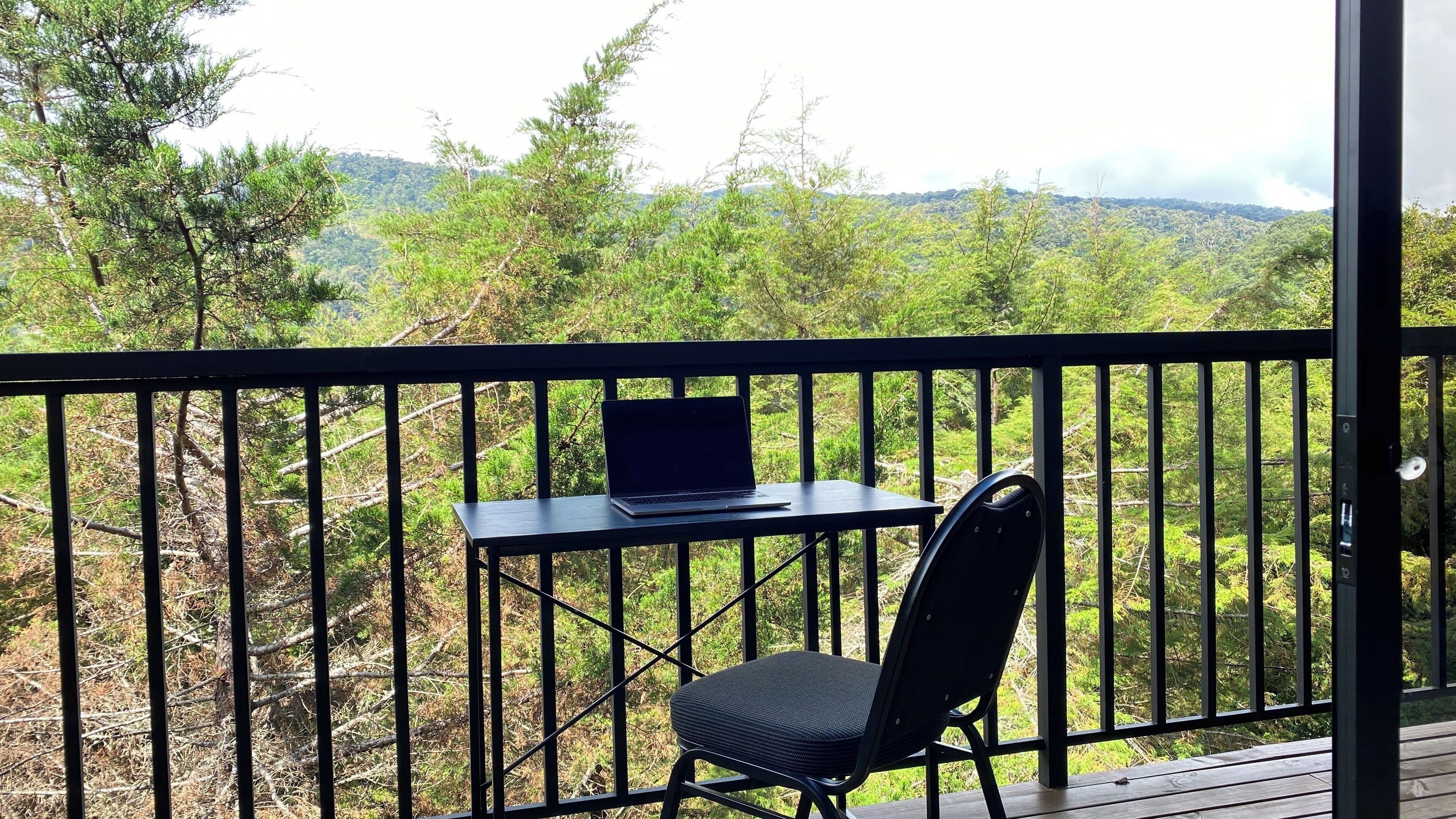 Un balcon avec une table, un ordinateur portable et une chaise, donnant sur un paysage boisé.