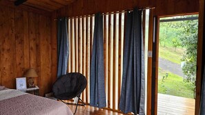 Ein Zimmer mit Holzboden, einem Bett, einem Sessel und Blick durch ein Fenster mit Vorhängen.