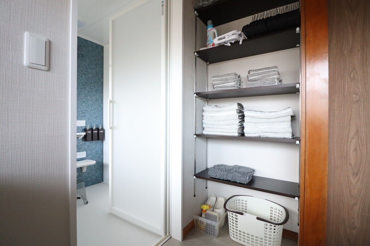 A bathroom with a blue tiled wall, a white door, and a shelf with folded towels and a basket.