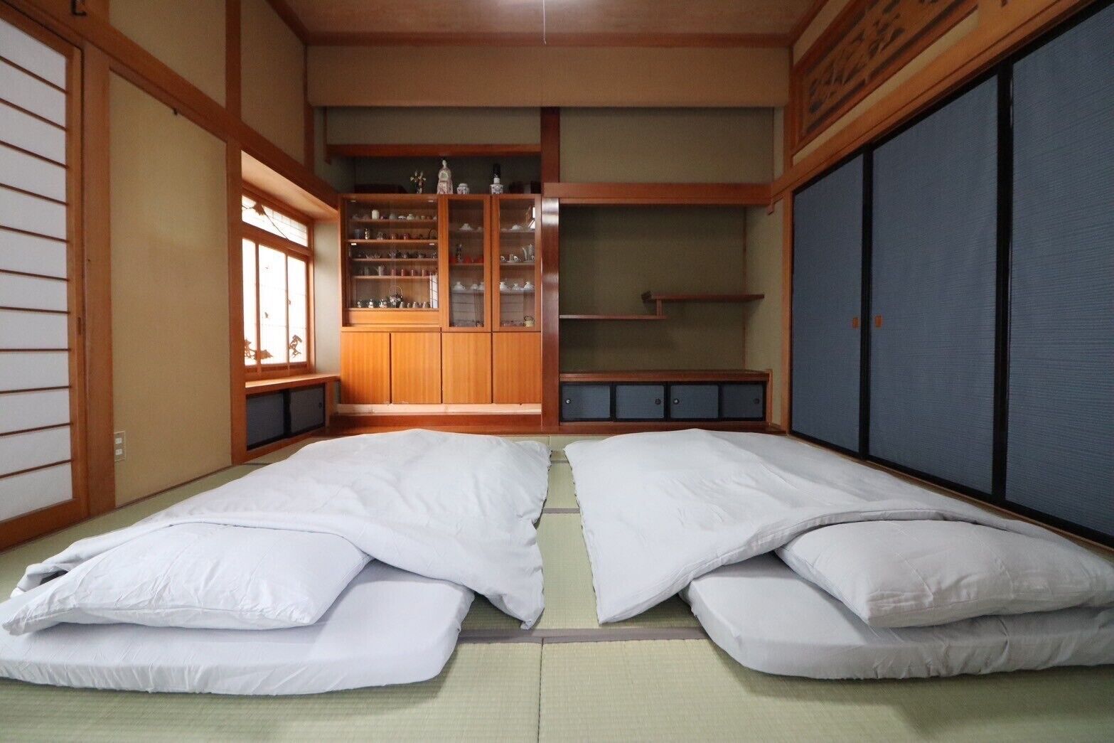 A traditional Japanese-style room with tatami flooring, a low wooden bed with white bedding, a wooden cabinet with glass doors, and sliding doors.