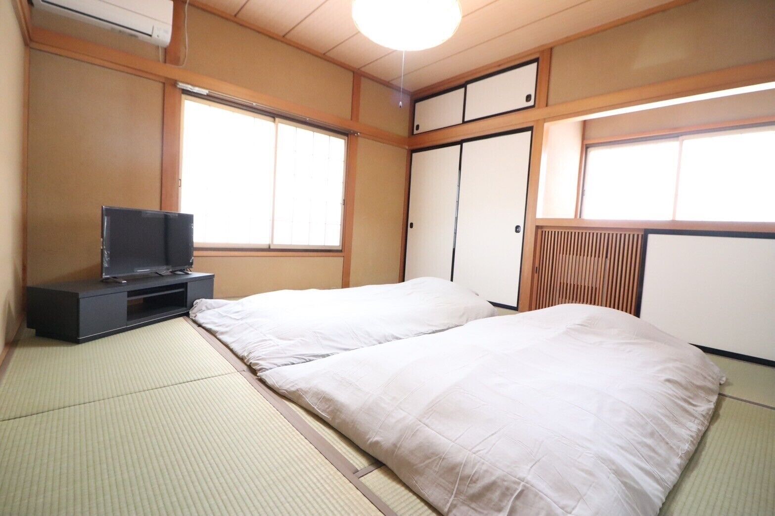 A traditional Japanese room with tatami flooring, a television on a low stand, two beds, sliding doors, and a ceiling fan.