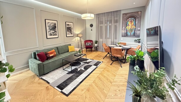 Ein modernes Wohnzimmer mit Sofa, Couchtisch und Essbereich. An den Wänden hängen Bilderrahmen, eine Chandelieber ist angebracht und es gibt ein Buntglasfenster.