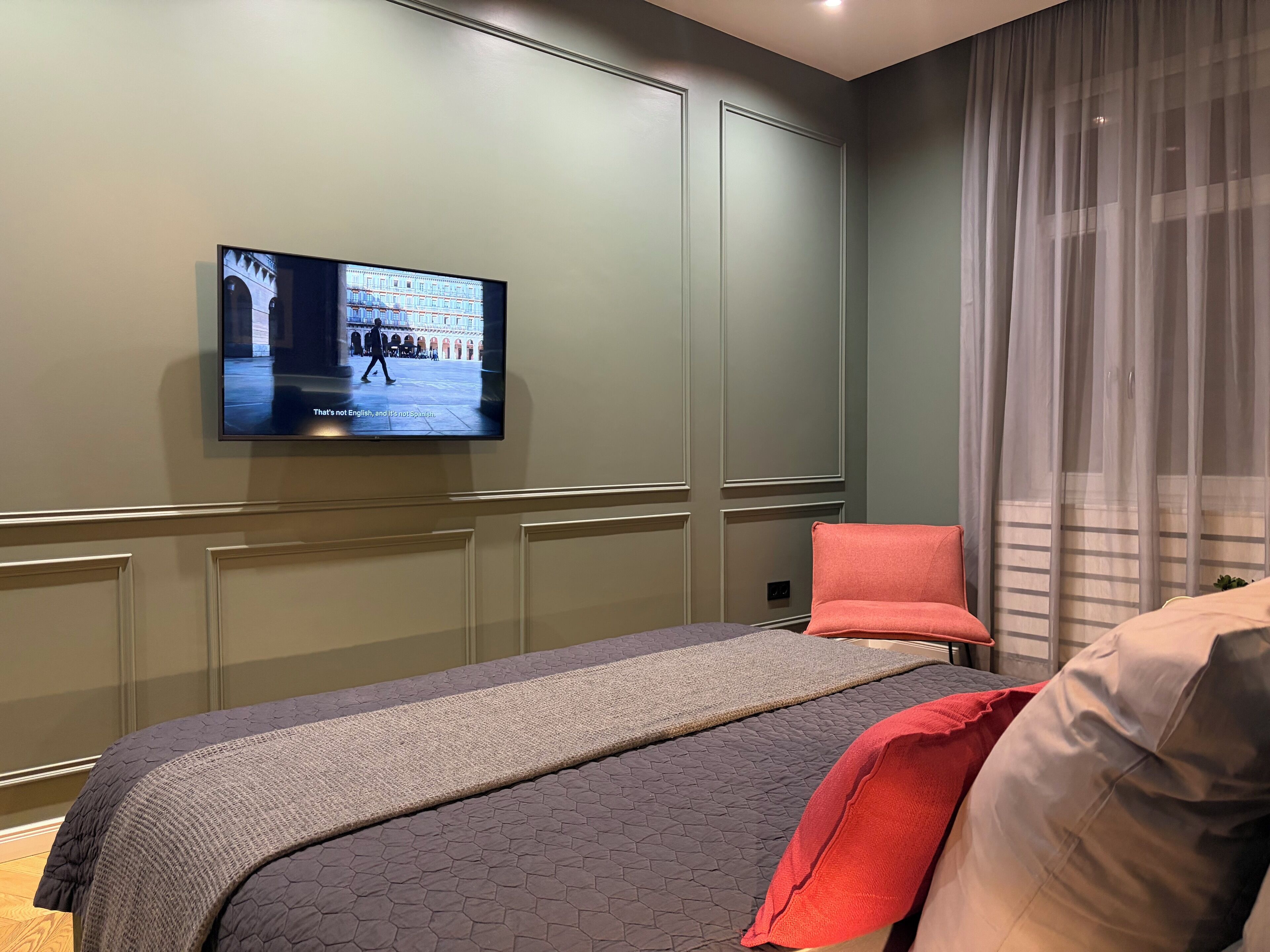 Camera d'albergo con un'ampia TV a schermo piatto montata a parete, un letto con coperta grigia e rossa, e una poltrona rossa.